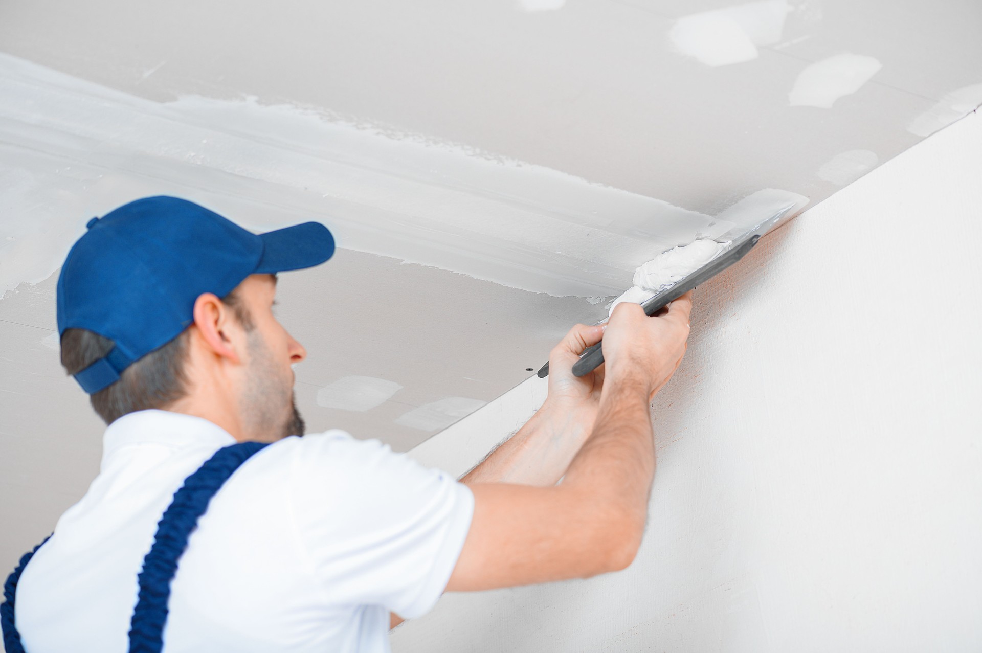 A master in uniform applies putty with a spatula to the reinforced joint of drywall on the ceiling.